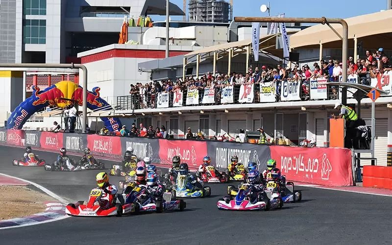Karting race in progress at the Dubai Kartdrome in Motor City, with a full grid of karts and spectators on the terrace above the pit lane